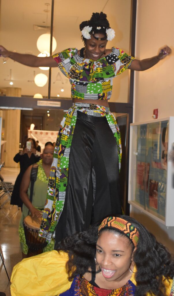Person wearing a colorful, patterned outfit and walking on stilts indoors.