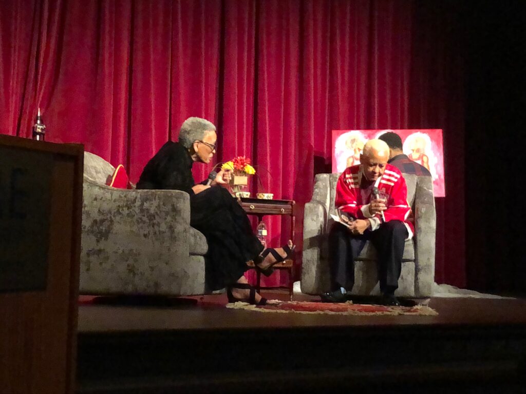 Two elderly women having a conversation on stage with a red curtain backdrop.