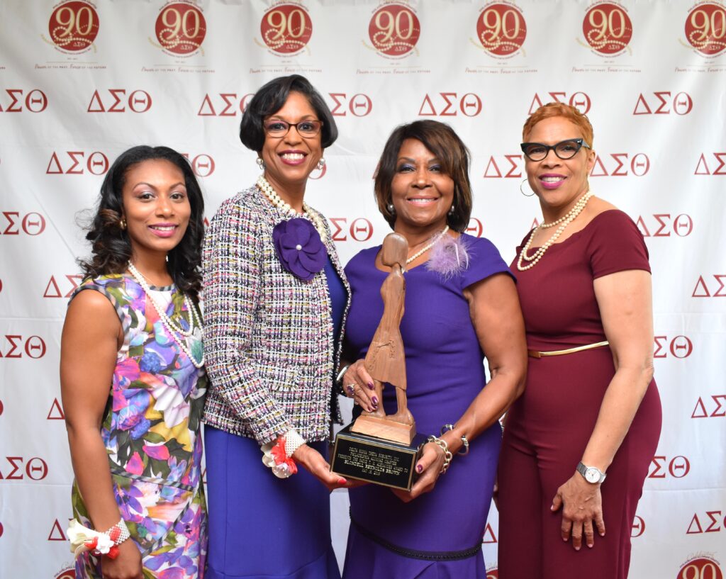 Four women posing together at a formal event with a backdrop of fraternity and sorority symbols.