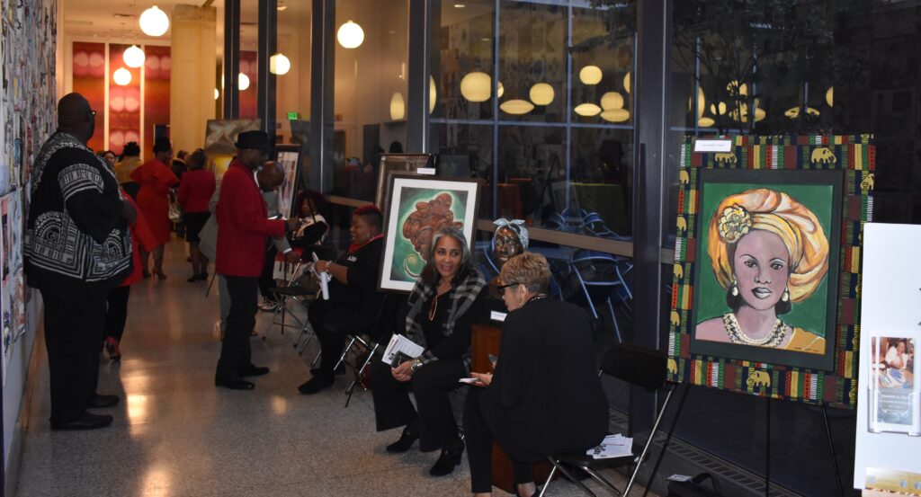 People seated and a man speaking indoors near framed portraits.