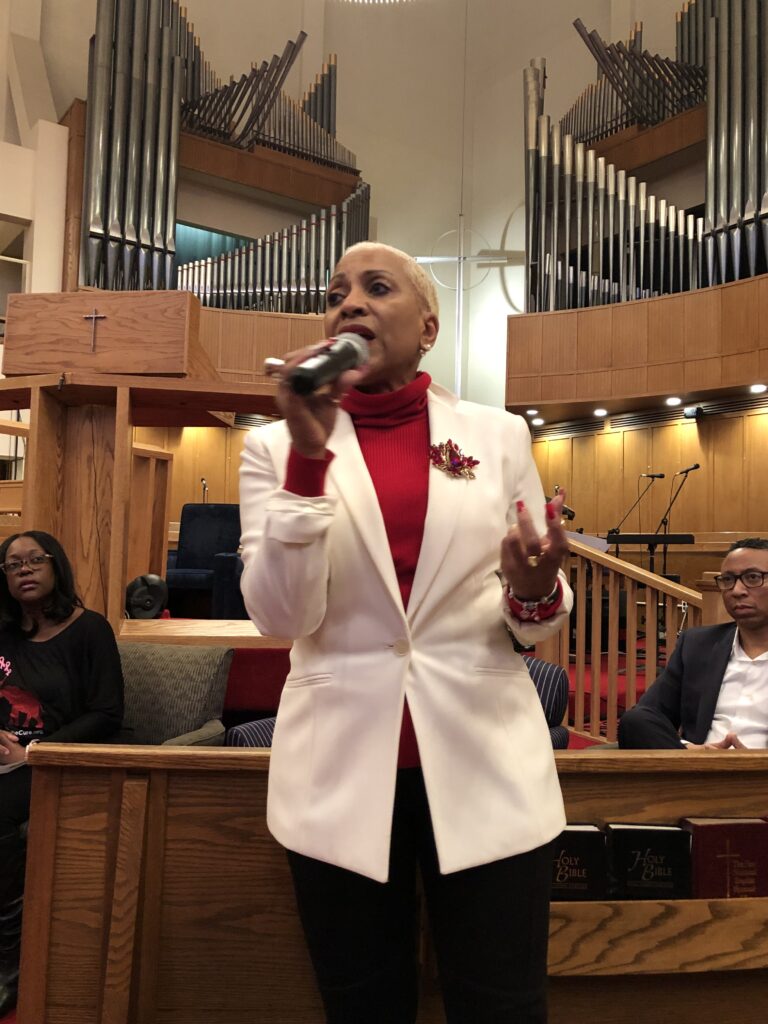 Woman speaking in a church, wearing a white blazer and red turtleneck.