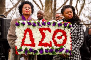 People holding a floral arrangement with Greek letters at a solemn event.