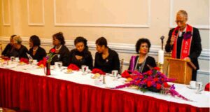 Women seated at a formal event table with microphones and flowers.