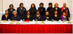 A group of eight women dressed in black posing behind a banquet table.