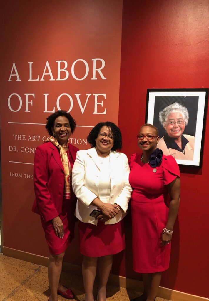 Three women smiling in front of a red exhibition wall with a portrait.