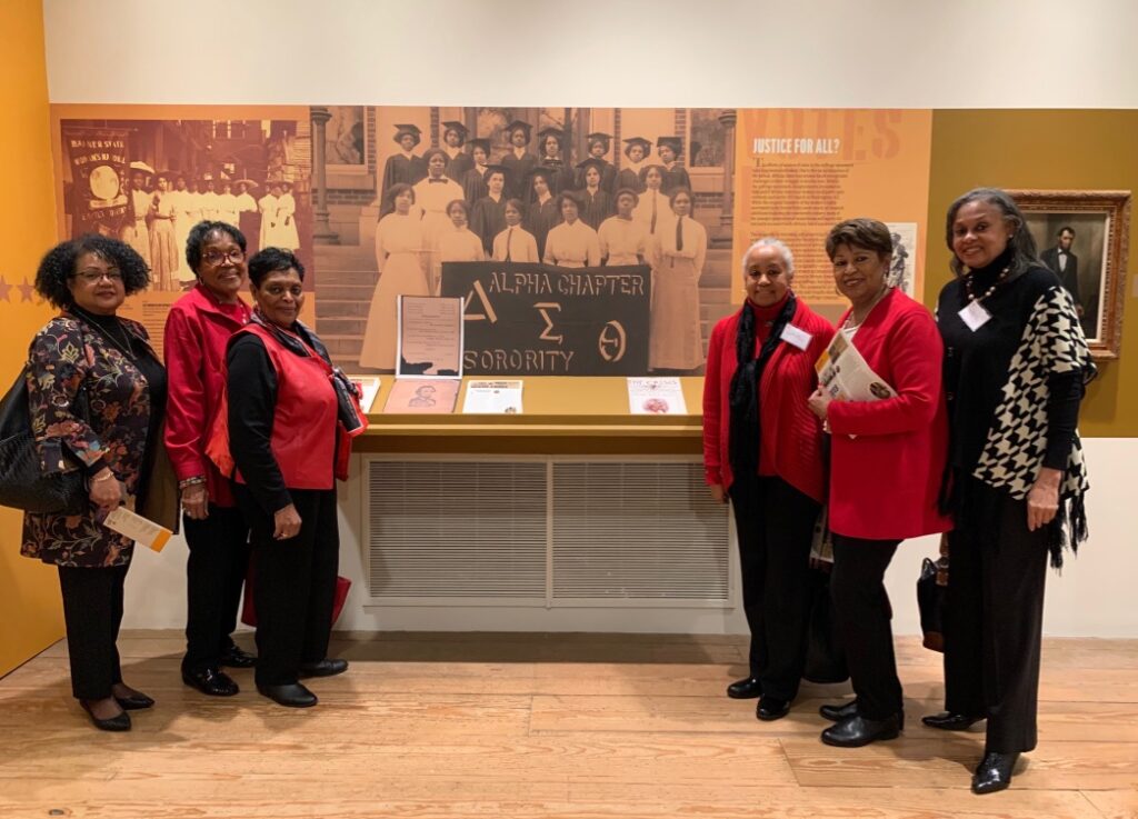 Four women in red jackets posing in front of a historical exhibit.