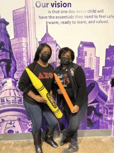 Two women holding oversized crayons, posing in front of a purple cityscape backdrop.
