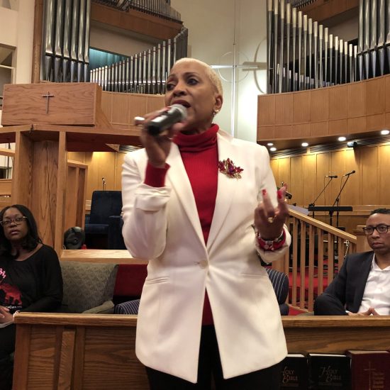Woman speaking in a church, wearing a white blazer and red turtleneck.
