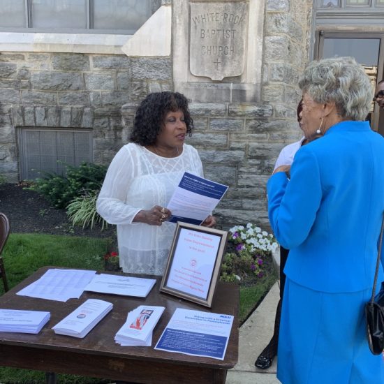 A woman in white speaking to a woman in blue at an outdoor table with documents.