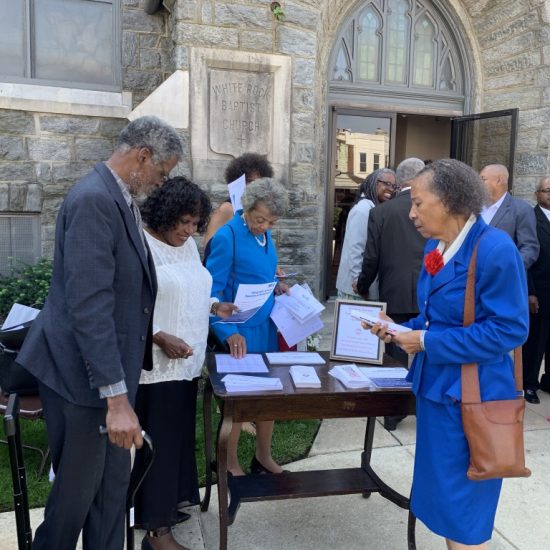 People gather at a table outside a stone building, exchanging documents.
