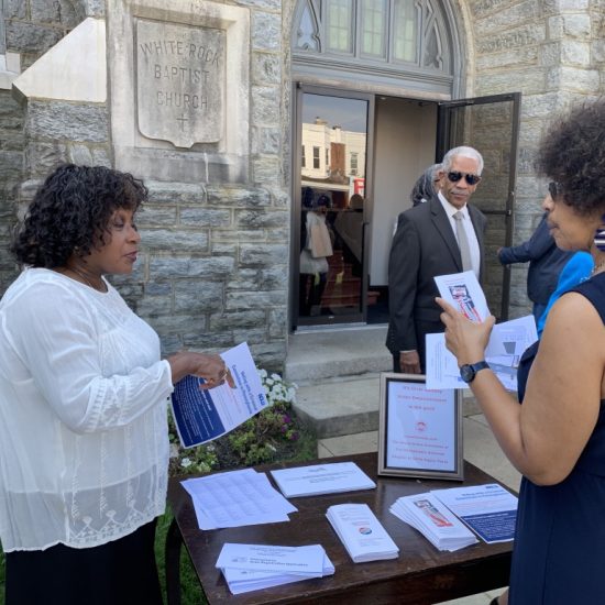 People gathered around a table with informational material outside a stone building.