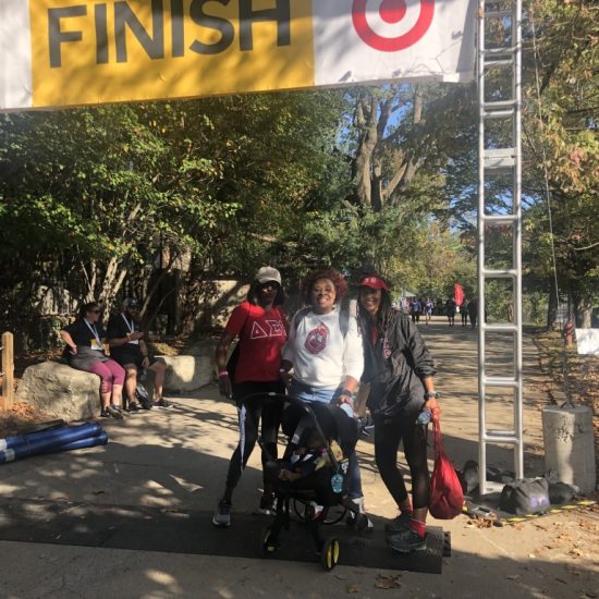 Three people with strollers at a race finish line on a sunny day.