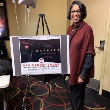A woman stands smiling beside a red carpet event sign.