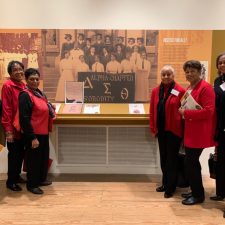 Four women in red jackets posing in front of a historical exhibit.
